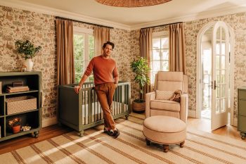 Nate Berkus standing beside a crib in a patterned nursery with a recliner, ottoman and bookcase